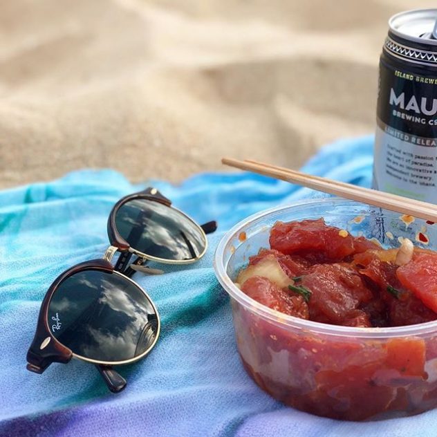 A photograph of Ray Ban sunglasses and a bowl of poke at the beach.